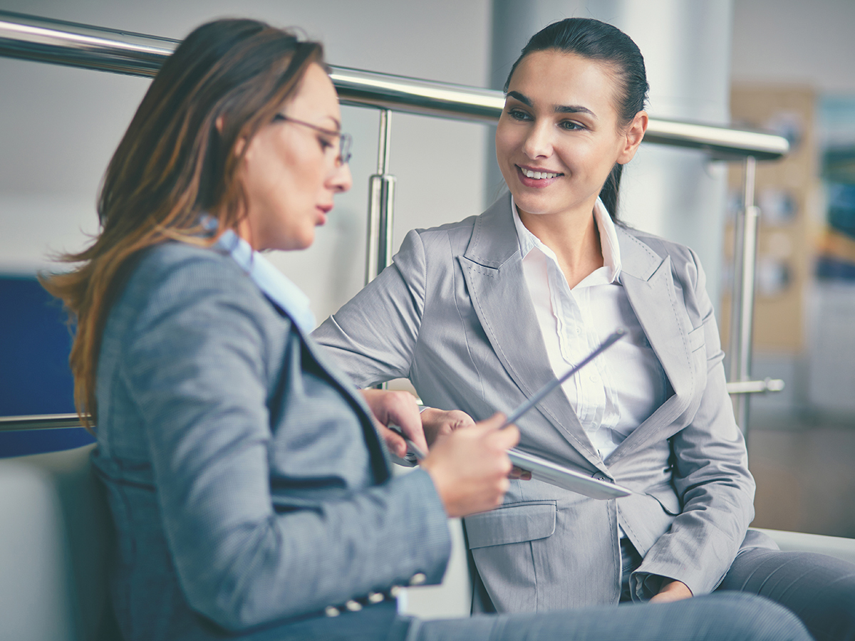 Two women in business attire talking and smiling while looking at a tablet
