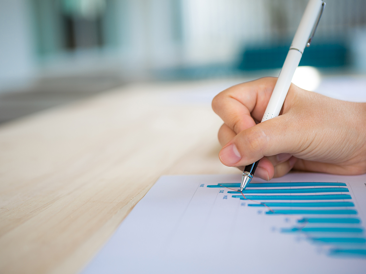 Hand holding a pen over a printed bar chart on a wooden desk
