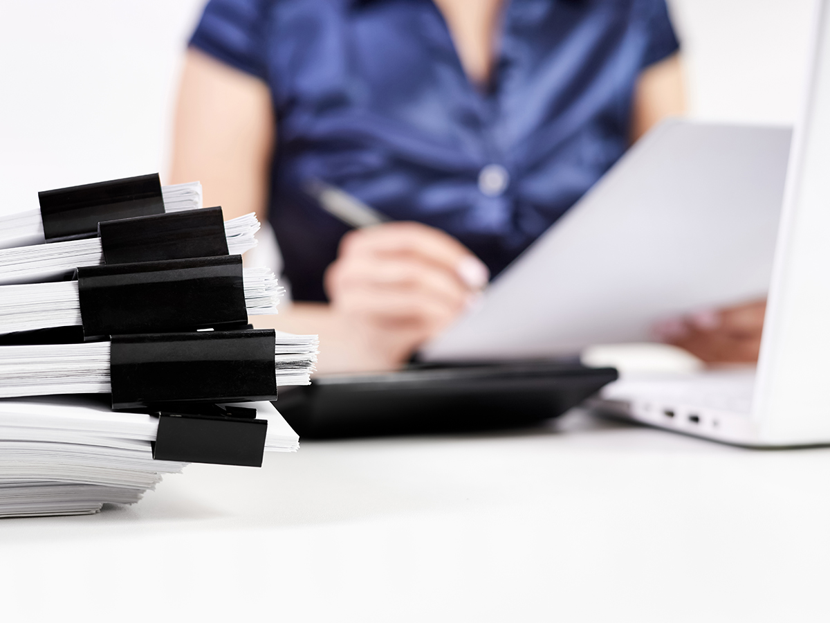 Stack of documents held with black binder clips on a desk, with a person working on papers in the background